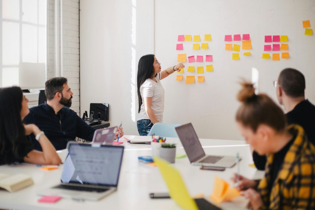 Woman placing colourful sticky notes on a wall in a meeting room with 4 other people sat around a table with laptops open.