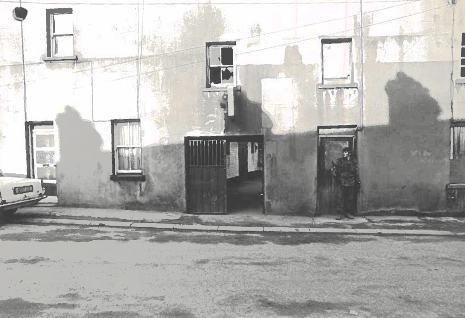 Black and white photograph showing the side entrance to Lyttle&rsquo;s Bar. At the lefthand side of the photograph is the tailgate of a light-coloured car. To the right of the photograph is a British Army solider. 