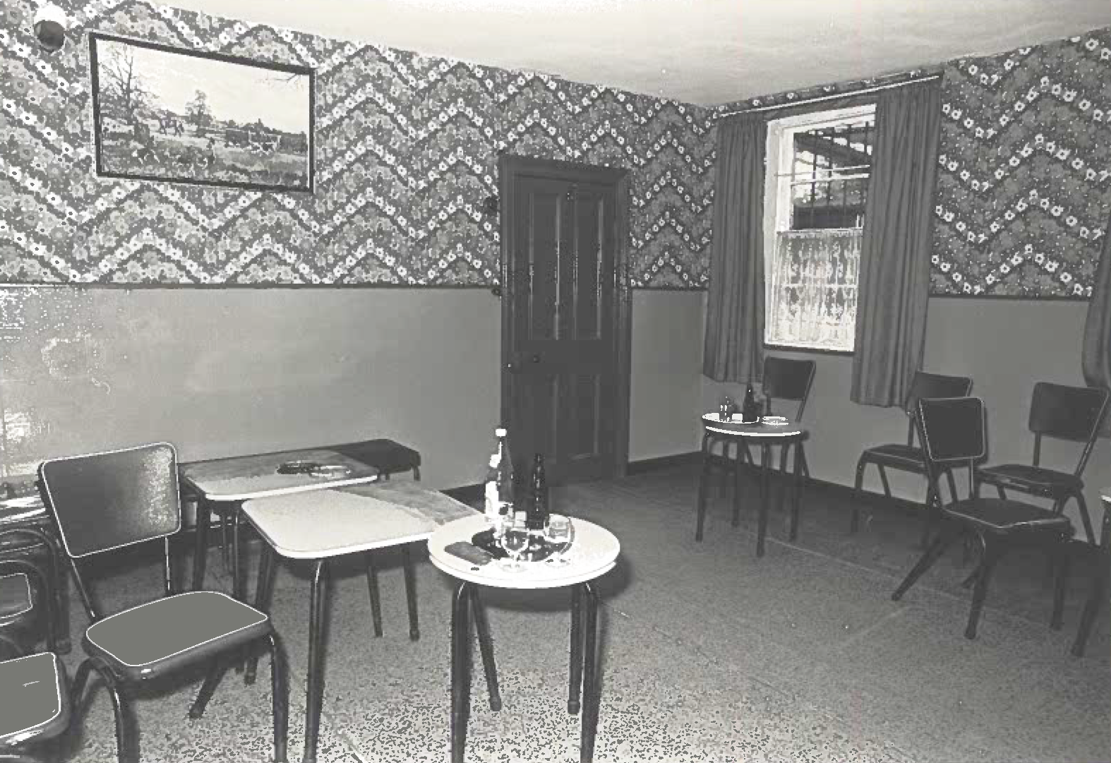 The photograph is black and white. At the centre in the foreground is a table with two bottles and two glasses. At the centre in the background is the door. Nearest the door is a table with a tray with glasses and a bottle on it. Tables and chairs are positioned throughout the room.   