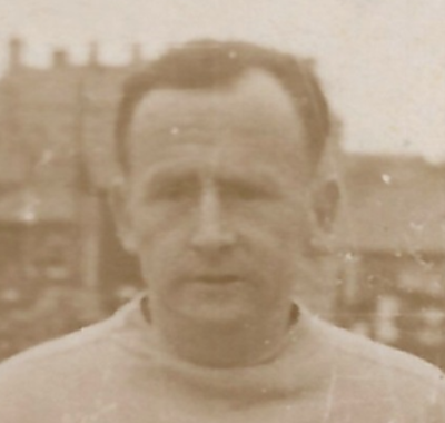 Head and shoulders photograph of Rory O&rsquo;Kelly. Rory is looking slightly to his right and is standing in front of a row of buildings. The photograph is in sepia tones. 