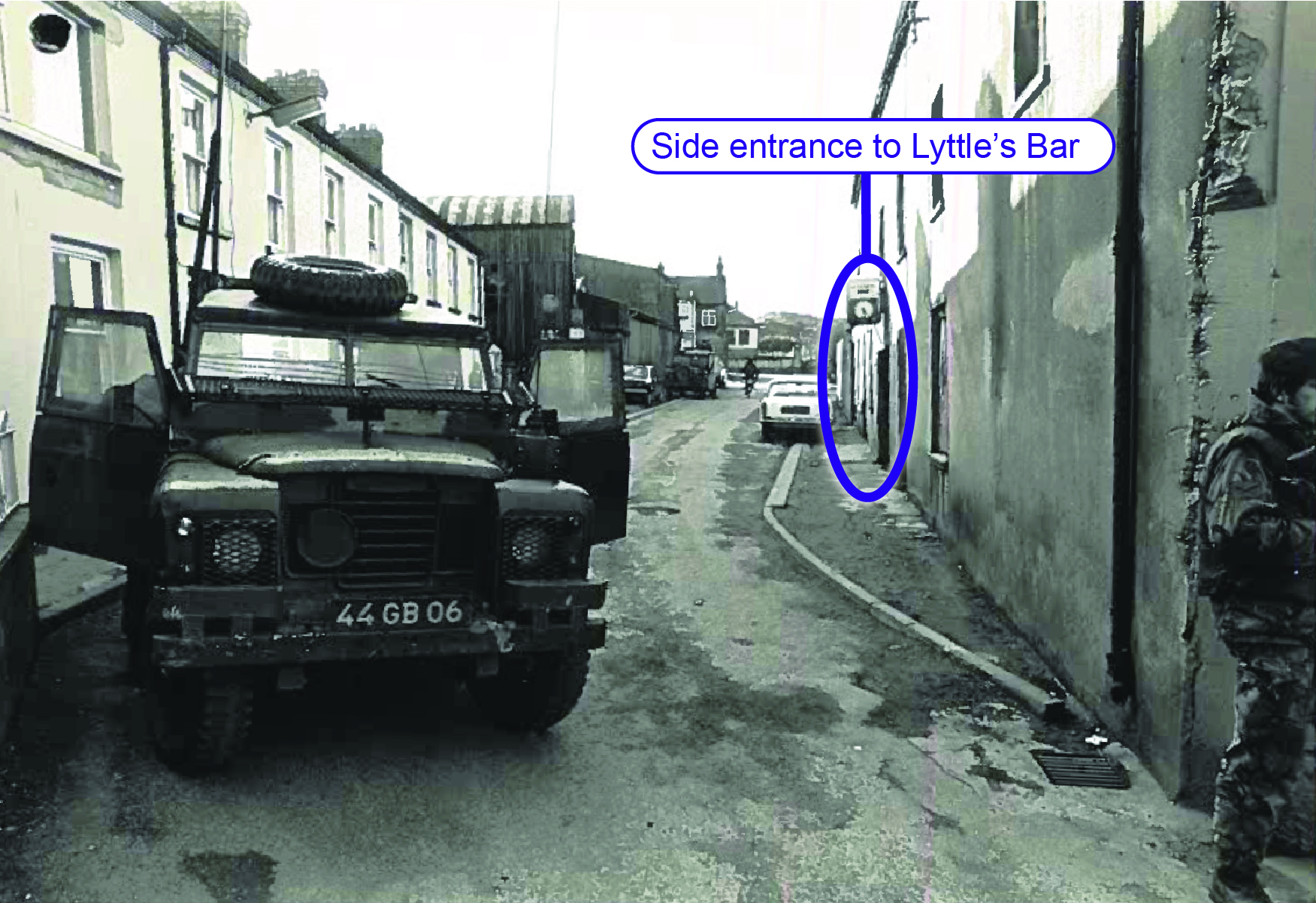 Black and white photograph showing the side entrance to Lyttle&rsquo;s Bar in the middle ground of the photograph. Labelling has been added by the Commission to highlight the entrance. In the foreground to the left is an armoured British Army vehicle. In the foreground on the right is a British soldier partly in frame. 