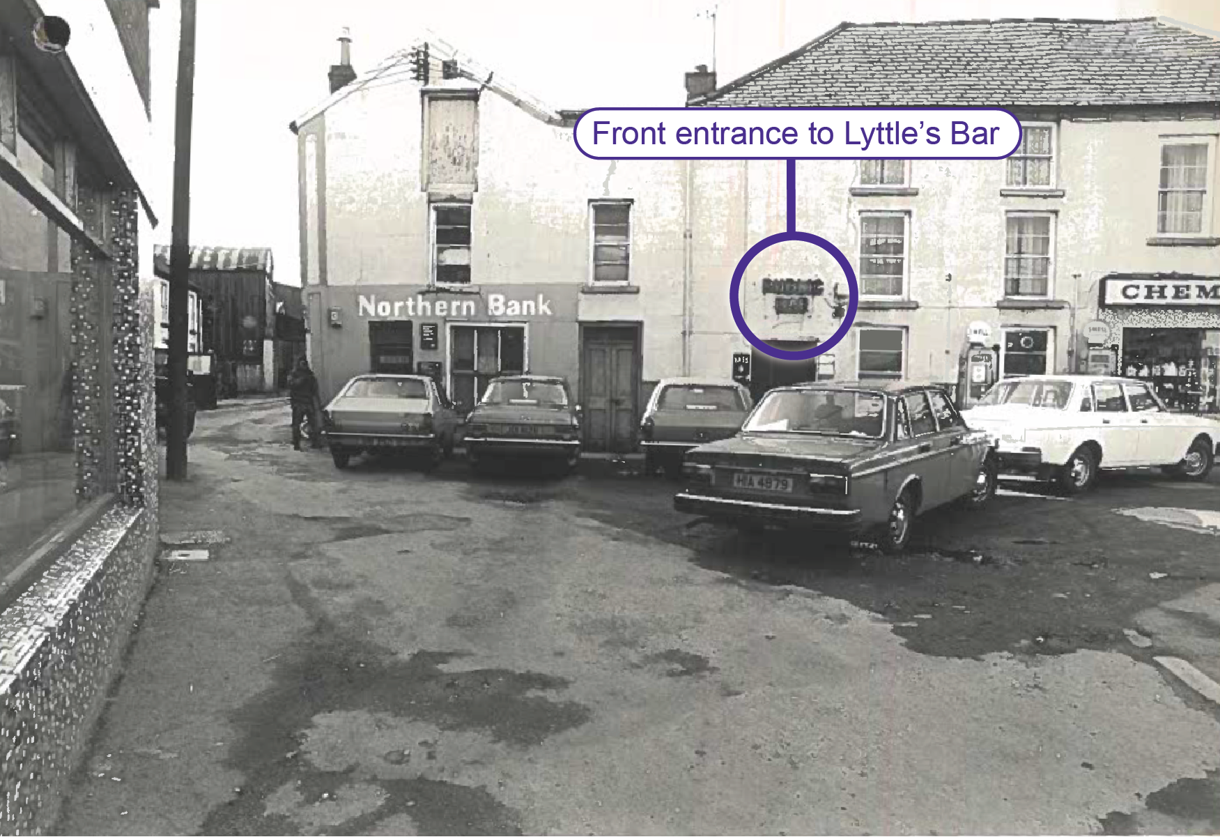 Black and white photograph showing the front of Lyttle&rsquo;s Bar. To the left of the bar is a conjoined unit displaying the signage &lsquo;Northern Bank&rsquo;. To the left of the frame at the corner of the front and side walls of the bank is a British Army soldier. To the right of the bar is a conjoined unit housing a chemist shop. The photograph displays only part of the signage for the shop. Five cars are shown in front of the row of buildings. Labelling has been added by the Commission to highlight the front entrance to Lyttle&rsquo;s Bar. 