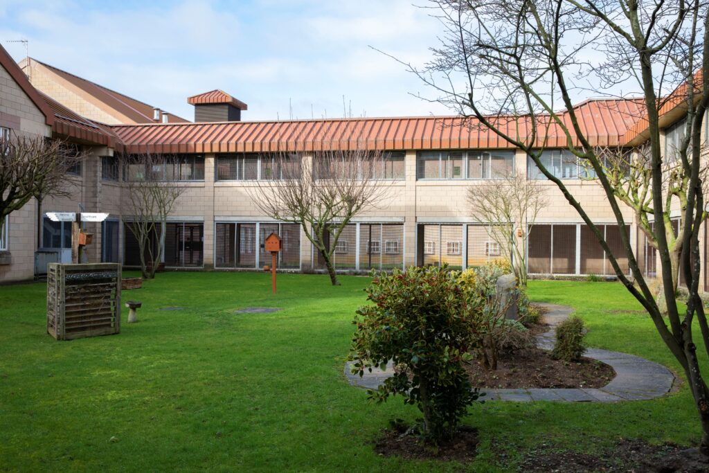 Inside HMP Bullingdon courtyard. There is a large brick building surrounding an outdoor space with green grass, trees and shrubs.