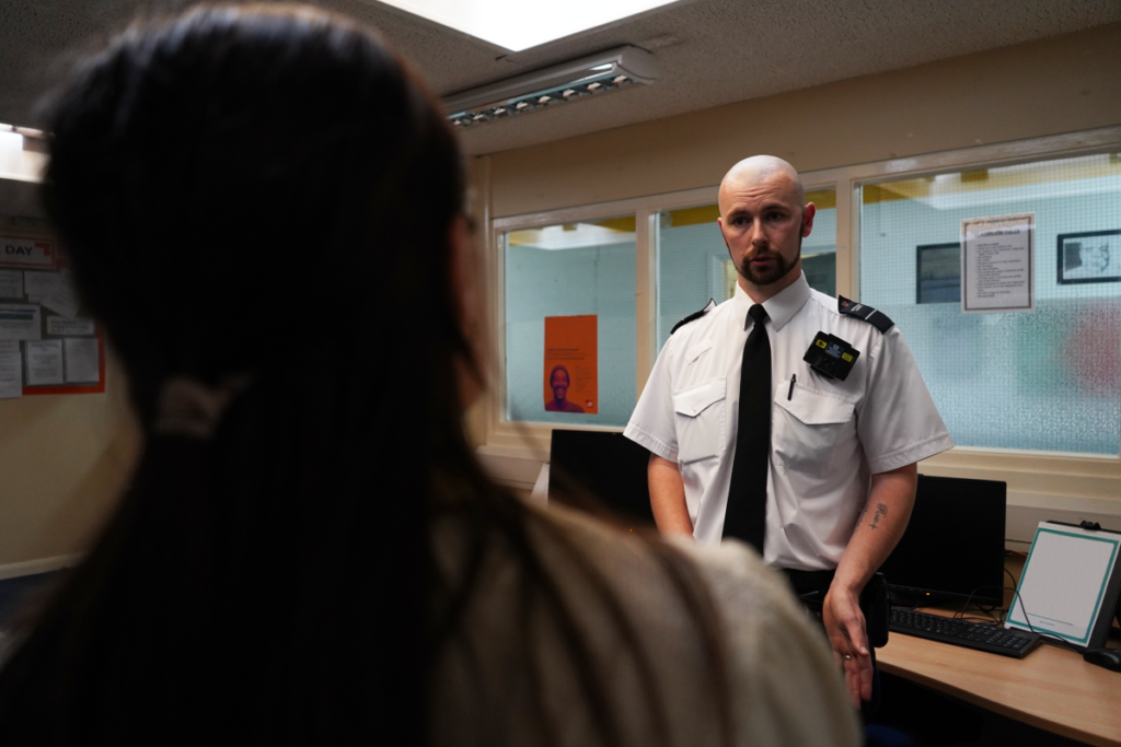A male prison officer standing inside an office talking to a female member of staff. The office has glass windows and there are computer monitors on desks.