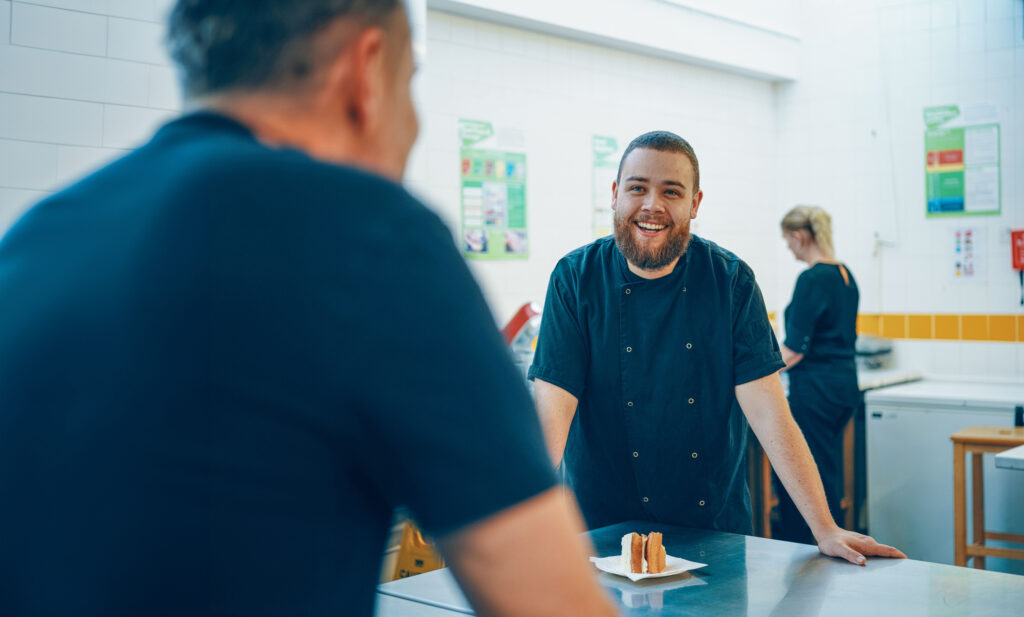 The image shows a cheerful man with a beard wearing a black chef’s uniform, leaning on a counter in a bright kitchen. He is engaging in conversation with another person whose back is to the camera. On the counter in front of him is a plate with a slice of cake. In the background, a woman in similar attire is working at another station, with posters and white tiled walls.