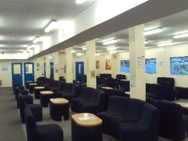 Photograph of the category B visits hall, a space with white walls and pillars, dark blue furniture and low coffee tables arranged in clusters.