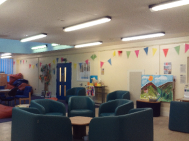 Photograph of the category C visits hall, a space with white walls and some decoration on the walls. Easy chairs are arranged around coffee tables.