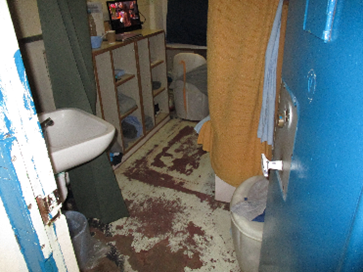 Photograph of a cell floor with lots of visible wear and damage. A sink is visible in the foreground and shelves can be seen in the background.