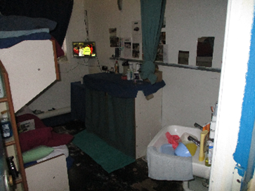Photograph of a cramped double cell with bunk beds, a sink and other furniture, as well as lots of the prisoners' personal effects, visible.