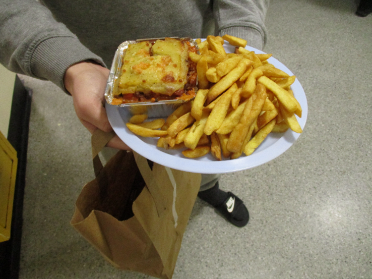 Photograph of a prisoner holding an evening meal. It consists of a generous portion of chips and a metal foil tray containing what appears to be a Shepherd's Pie.