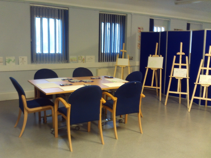Photograph of the L wing recreation area. Four artists' easels stand to one side, while in the middle there is a square table with chairs arranged around it. Art can be seen on the table.