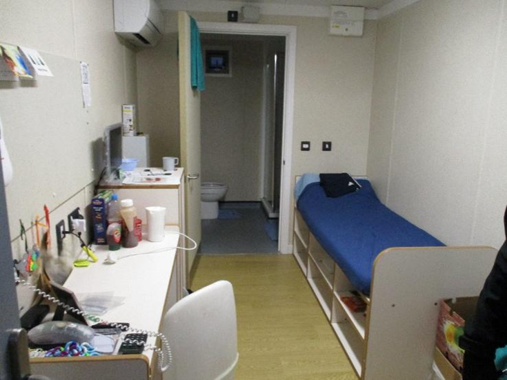 Photograph of a cell on M wing, featuring a single bed to one side and a desk and other furniture on the other side. The occupants' belongings are arranged on the desk.
