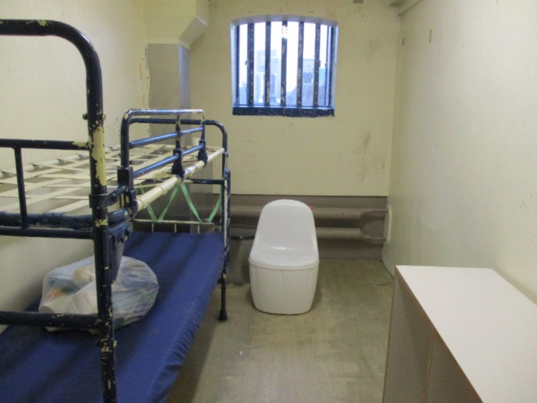 Photograph of a first night cell, with a bed on one side, a plastic moulded chair underneath the window and a desk to one side.