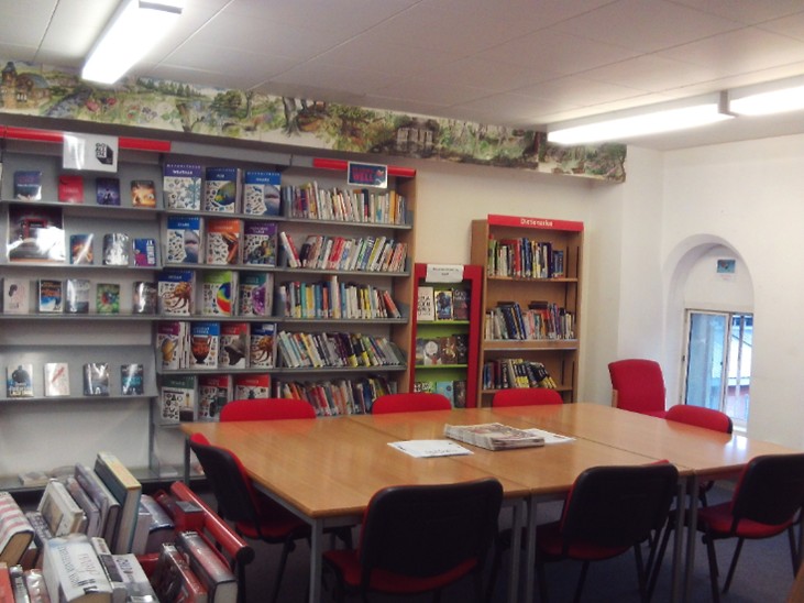 Photograph of the library, featuring shelves of books against one wall and a table with several chairs around it.