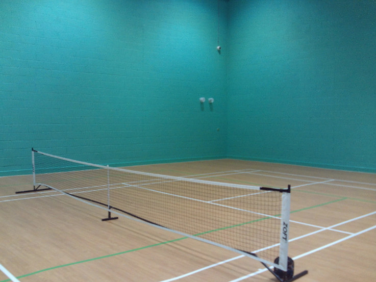 Photograph of the sports hall. The walls are painted aquamarine and the floor is of light-coloured wood. The floor is marked for various sports and a tennis net has been erected.