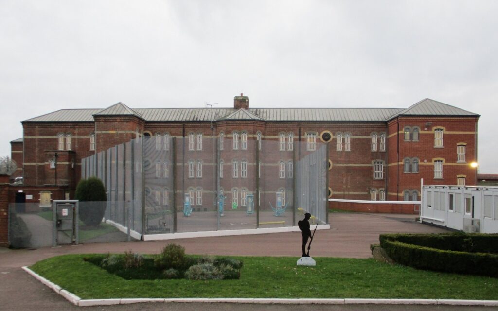 Photograph of the exterior of HMP Aylesbury. An area with grass and bushes can be seen in the foreground, with a fenced off yard and a three-storey brick building them. Part of a portakabin is visible at the right of the image. On a small grass area at the bottom of the image a silhouette statue of a soldier holding a rifle in one hand and with his head bowed can be seen.