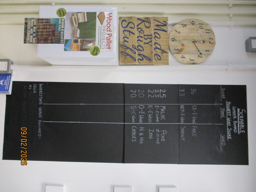 Photograph of the Scrabble leader board in the library. The floor-to-ceiling chalk board has columns with scores and prisoner nicknames on it.