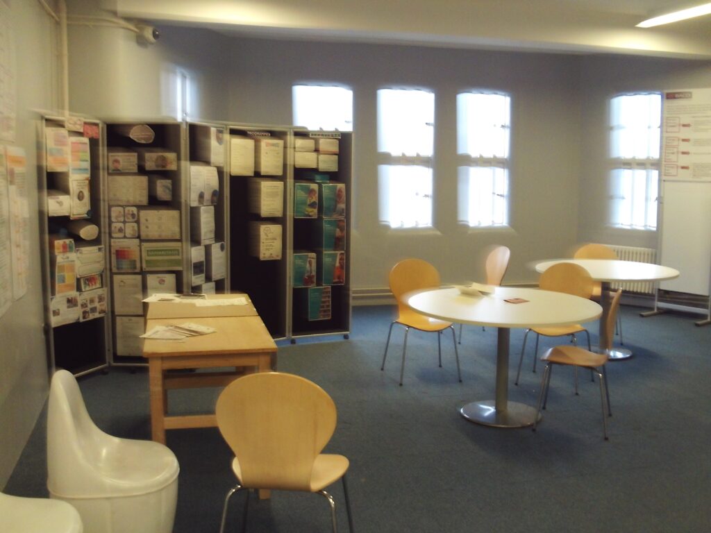 Photograph of the induction room on G wing. The space has several tables with chairs arranged around them, and to one side there is a tall display board with lots of information displayed on it.