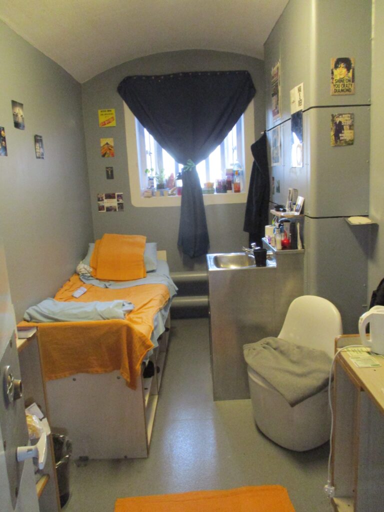 Photograph of a cell with a prisoner's belongings in it. Postcards decorate the walls and personal effects are arrnged on the furniture and around the wash basin.