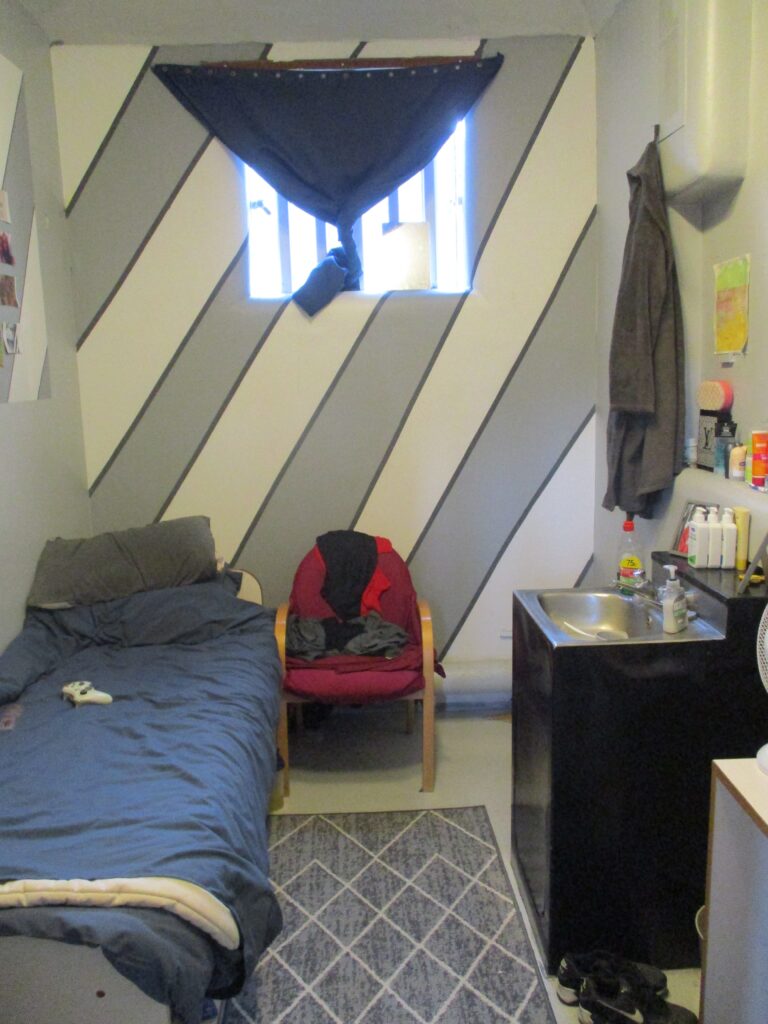Photograph of an occupied cell. There is grey and white striped wallpaper on one wall, a rug on the floor next to the bed, a chair with some clothes on it and personal effects arranged the wash basin.
