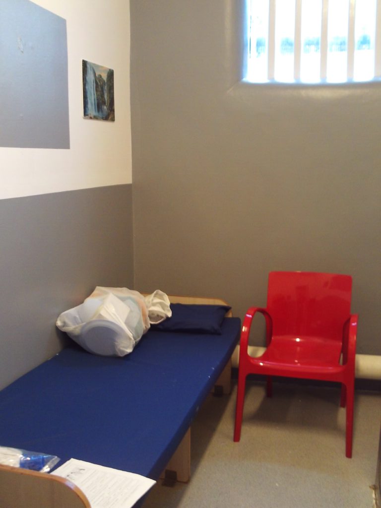 Photograph of a cell prepared for a new arrival. The bed has a mattress on it, with bedding and other things the occupant will need in a bag. A red chair is positioned next to the bed.