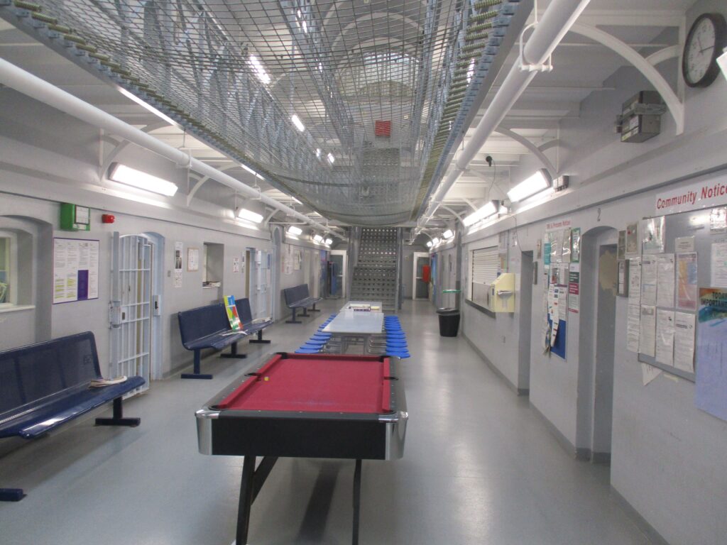 Photograph of a landing. Cell doors can be seen off to both sides, benches are arranged down one wall, and there are pool tables down the middle of the space.