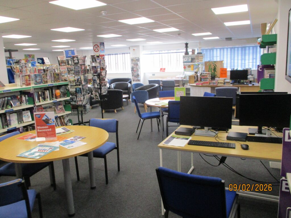 Photograph of the interior of the library, showing a seating area with both circular tables with chairs arranged around them, and desks with computer terminals on them visible.