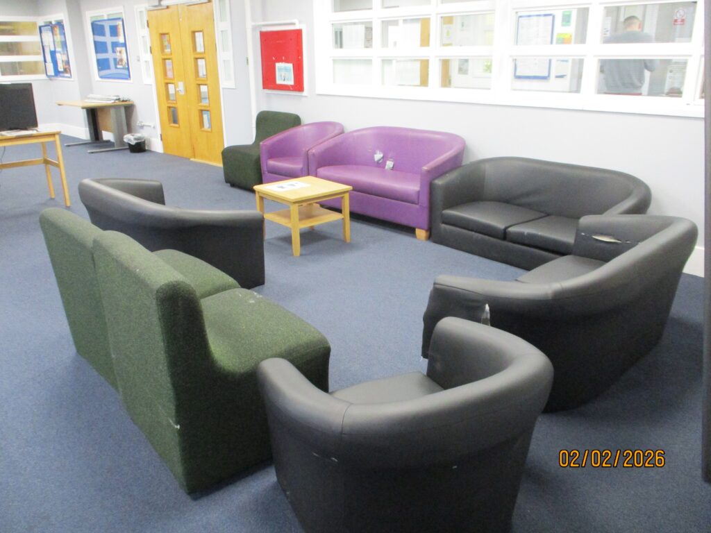 Photograph of the seating area in reception for holding prisoners and first night interviews. Easy chairs are arranged around a small coffee table. The chairs are upholstered in a variety of colours. Beyond the seating there is a set of yellow doors and a table with a screen on it.