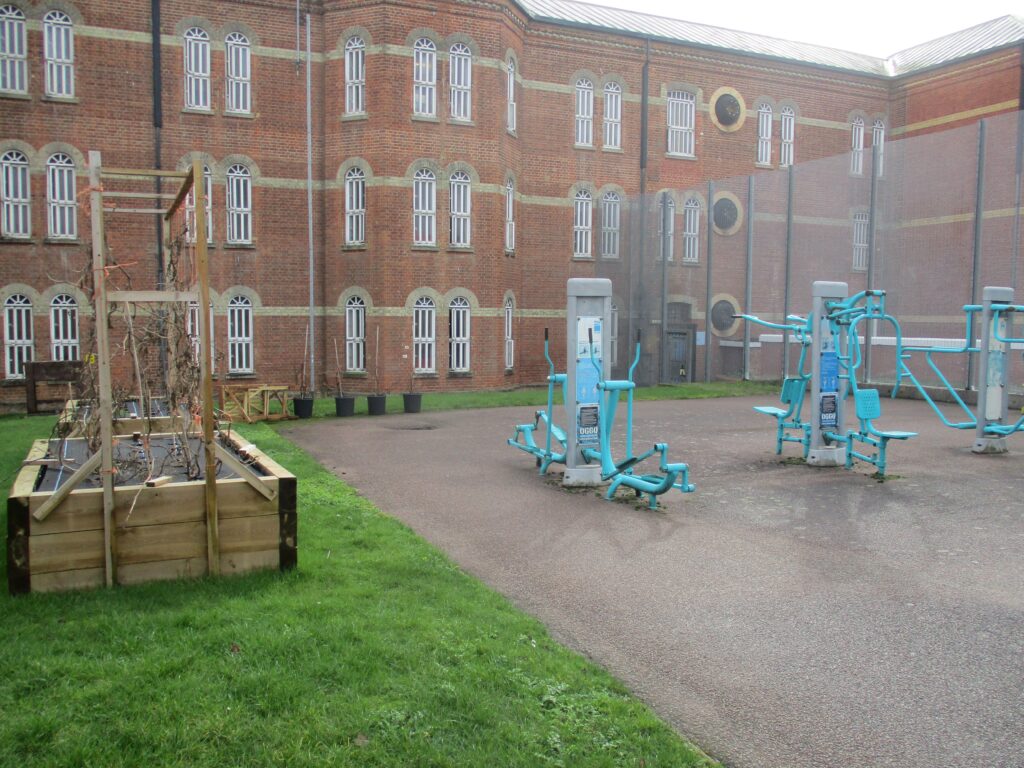 Photograph of an exercise yard. To one side there is a concreted area on which there are some pieces of exercise equipment, to the other there is a grass area with a raised bed planter in the middle of it. A three-storey brick building sits behind the yard.