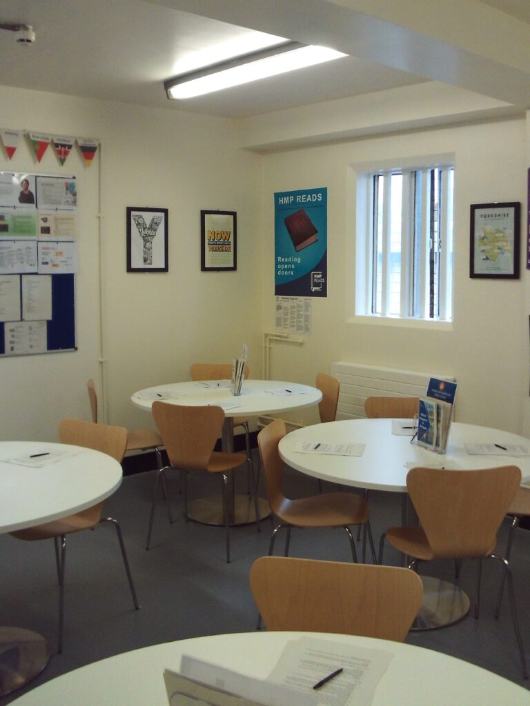 Photograph of the induction classroom. There are four tables with chairs arranged around them. There is literature on the table and lots of information posters are displayed on a noticeboard and on the walls. A window to one side of the room lets natural light in.