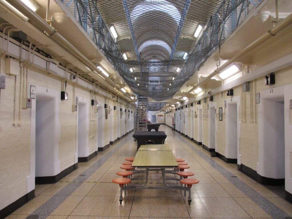 Photograph of the interior of a wing at HMP Leeds. It shows a long communal space with tables and stools, a pool table and a table tennis table arranged down the centre. Doorways into cells can be seen down both sides of the space, and there is netting hanging above it, through which the upper landing can be seen.