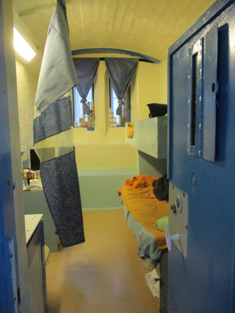 Photograph of a cell that was being shared, but was designed for one. The photograph has been taken through the doorway, with the door open. There is a double window at the back of the cell and some furniture is visible to the other side.