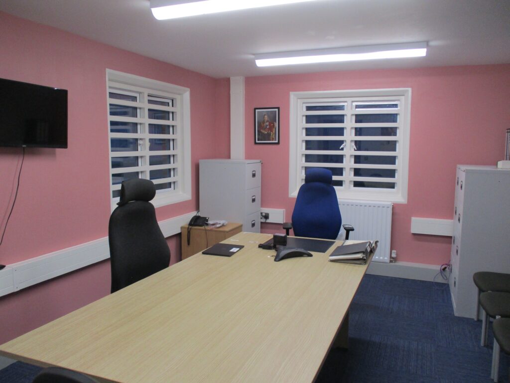 Photograph of the adjudication area, a room with walls painted pink. In the centre of the room there is a table with two chairs tucked underneath it. Other furniture in the room includes filing cabinets and wall-mounted screen.
