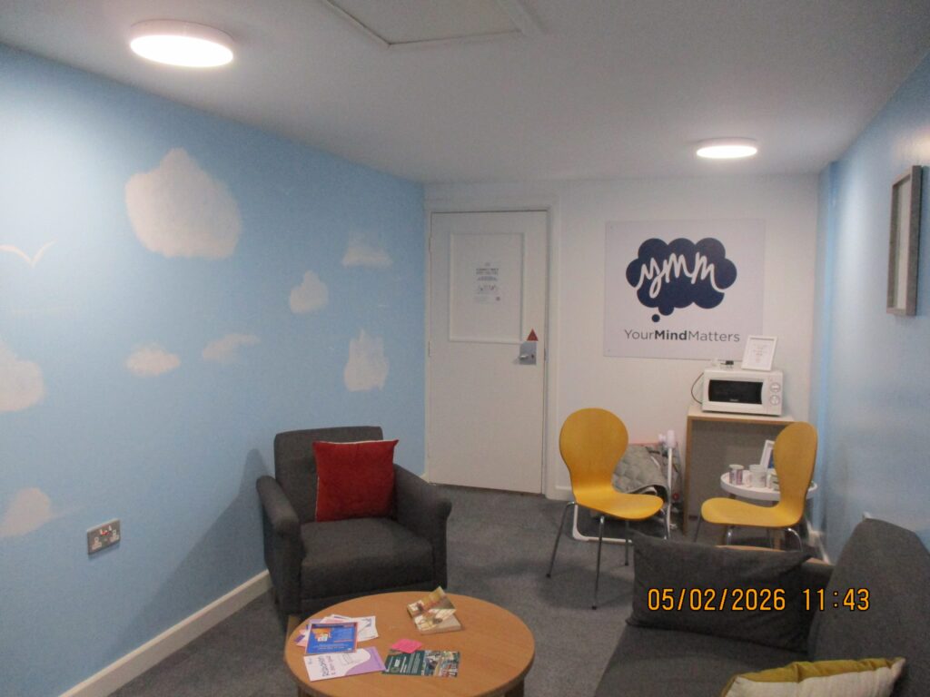 Photograph of the baby bonding session room. Easy chairs are arranged around a low table, which has some leaflets on it. One of the walls has been painted with a floor-to-ceiling mural of clouds in a blue sky.