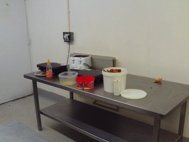 Photograph of cooking facilities on a residential unit. A stainless steel table is positioned against a wall. On its surface can be seen equipment including a toaster and some Tupperware containers.