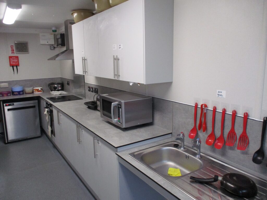 Photograph of a kitchen on the pods, with food preparation equipment visible on the long work surface. Above it there are wall-mounted cupboards.