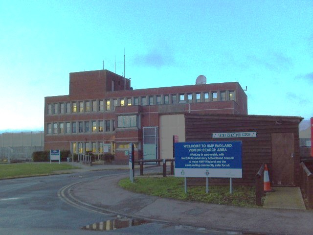 Photograph of the exterior of HMP Wayland in which a three-story red brick building can be seen in the background, with a smaller brick building and a sign with the prison's name on it in the foreground.