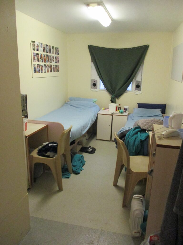 Picture of a double cell on a new unit. A bed is arranged against each of the side walls, with a chair and desk next to each. There is a window in the back wall with a curtain draped across it.