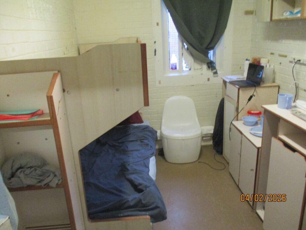 Photograph of a double cell on the old unit, featuring bunk beds to one side, and furniture including a chair and cupboards to the other.