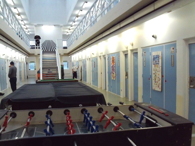 Photograph of the induction unit, with a view down the central area in which there is a table football table and pool table, with a staircase leading to the upper landing in the background. Cell doors can be seen on both sides of the image.