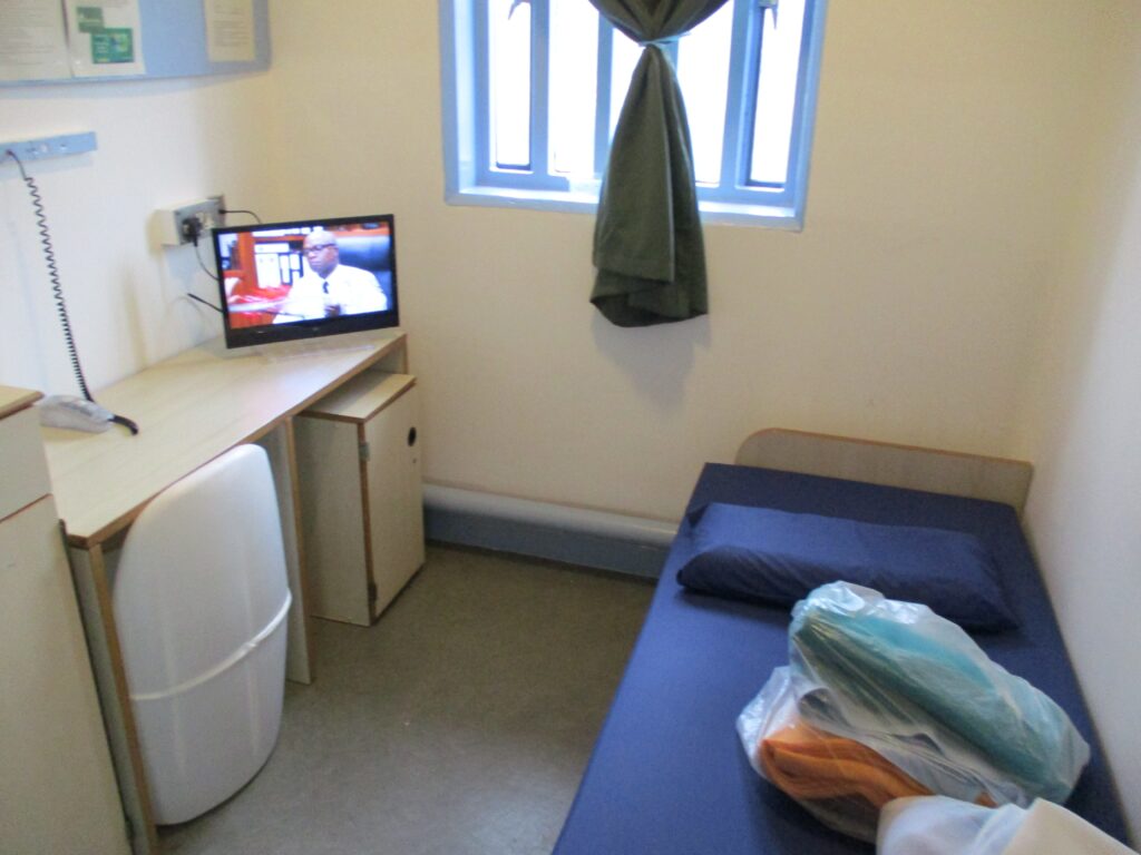 Photograph of a new induction cell. There is a bed to one side, and a desk and chair to the other. There is a television at one end of the desk.