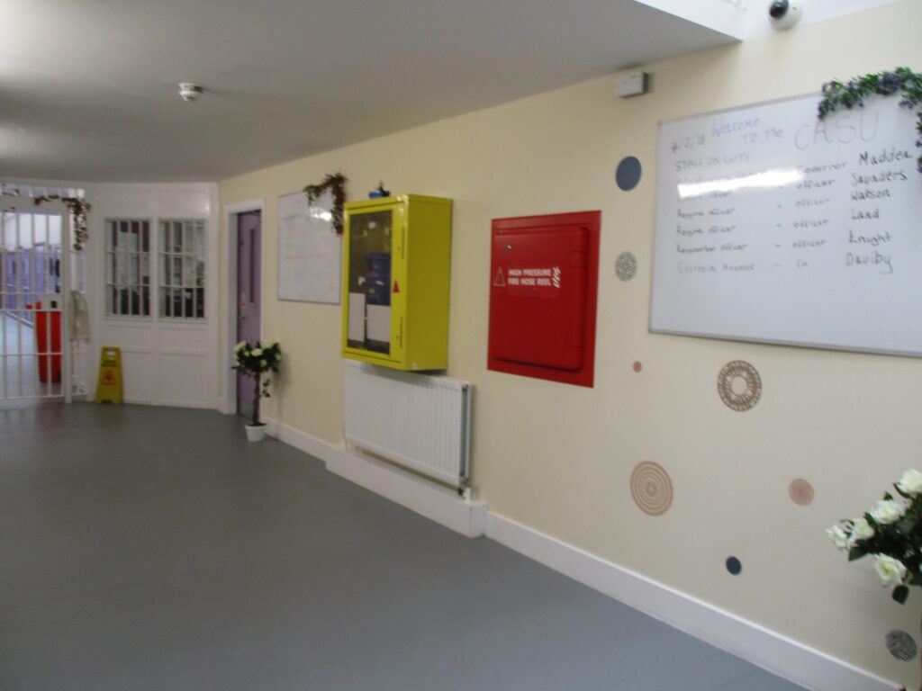 Photograph of a corridor in the segregation unit. There is a wall mounted fire hose and other displays of information can be seen.