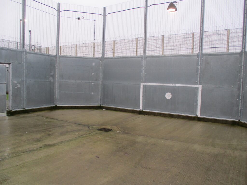 Photograph of the segregation unit exercise yard. It has a high fence and there is a football goal painted on the visible end of the yard.