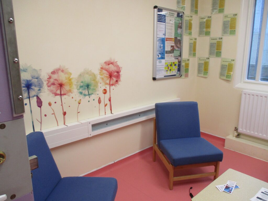 Photograph of part of the segregation unit library. Two easy chairs are arranged around a coffee table, there is a mural painted on one wall, and displays of information are mounted on the wall.
