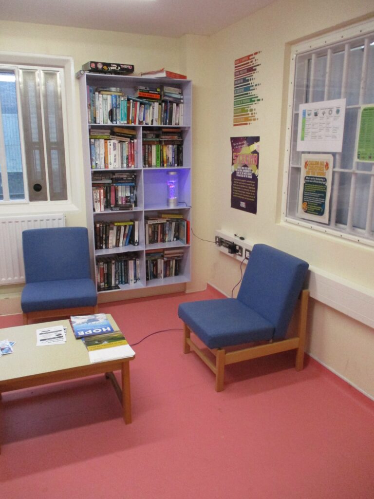 Photograph of another area of the segregation unit library. There is a tall bookcase in one corner and in front of it are a coffee table wand easy chairs.