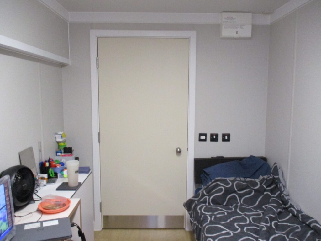 Photograph of a single room in a pod.  There is a bed with a blue and white duvet cover to one side, with a desk on which the occupants' things are visible on the other.