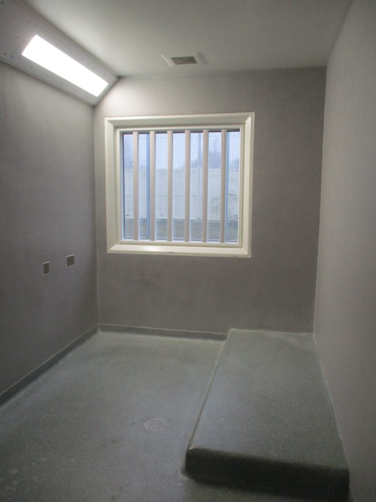 Photograph of a special accommodation cell. The walls and floor are of grey concrete and there is a low bed to one side, with a barred window in the rear wall.