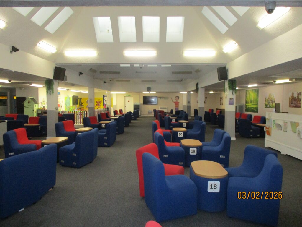 Photograph of the visits hall, with tables arranged in two rows with chairs around them. The chairs are blue or red. Skylights illuminate the room.