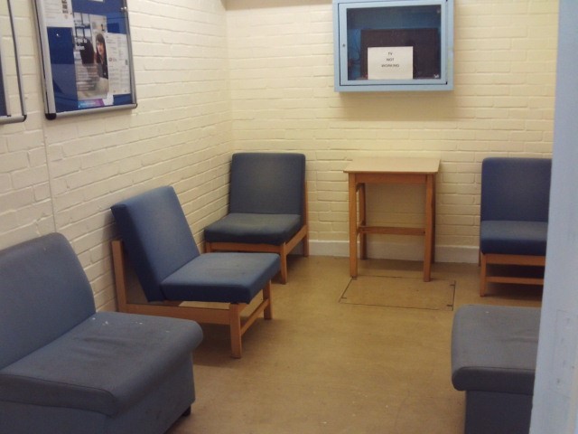 Photograph of the waiting room in reception. The room has walls painted white and some blue-upholsthered easy chairs. There is a small table against one wall and some notice boards on the walls.