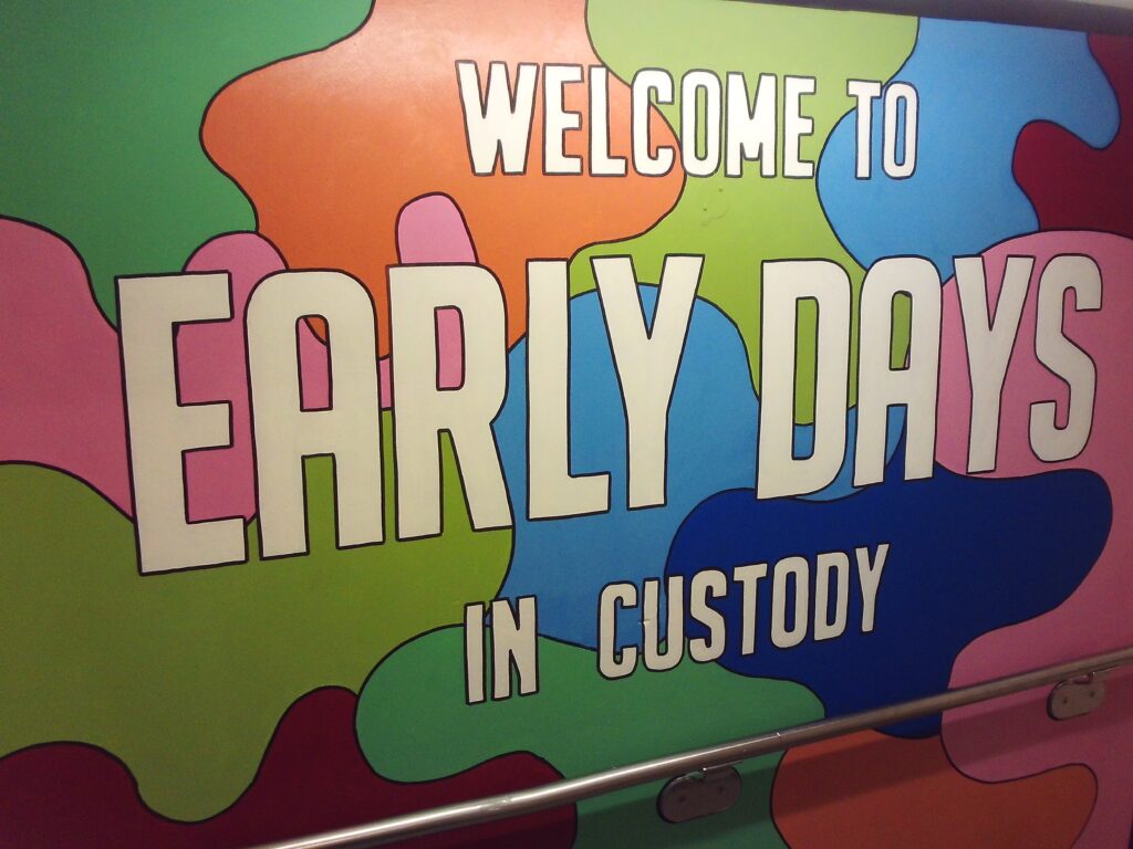 Photograph of a mural with the words 'Welcome to early days in custody' on it. The letters are painted in a cream colour with black outline around them; the background is a mixture of bright colours including orange, green, blue, red and purple.